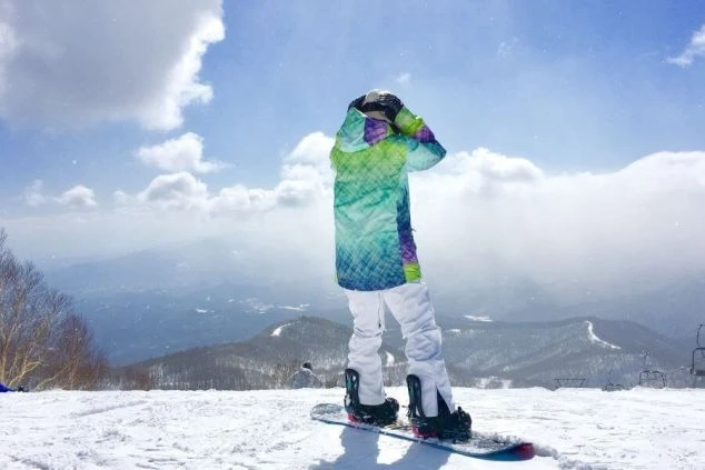 雪山ゲレンデで、スノーボードを持った男女グループが笑顔で座っている集合写真。リゾートバイトの休日に仲間とスノーボードを楽しむ様子。