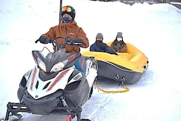 雪原でスノーモービルがゴムボートを牽引している画像。スノーモービルを運転する人と、ボートに乗って楽しむ2人の姿があり、雪上アクティビティを楽しむイメージ。