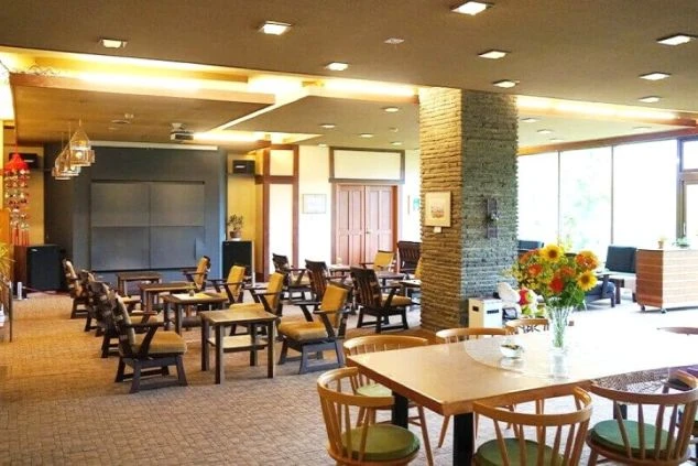 白馬にあるホテルのロビーの画像。広々とした空間にテーブルと椅子が多数配置され、暖炉のような石積みの柱や花瓶のひまわりが温かい雰囲気を演出する。リゾートバイトの職場となる施設の雰囲気。