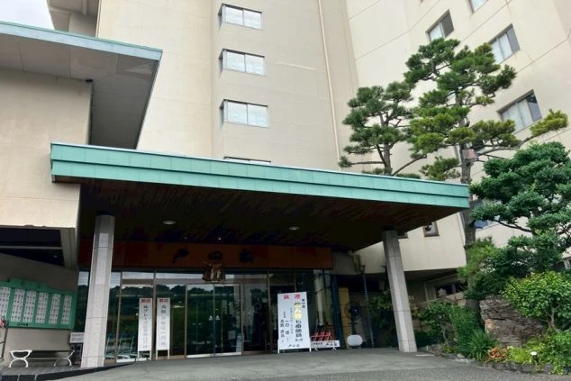 大規模な旅館またはホテルのエントランス。大きな庇の下にガラスの自動ドアがあり、旅館の名前が書かれた看板や案内が見える。建物の側壁には立派な松の木が植えられている。