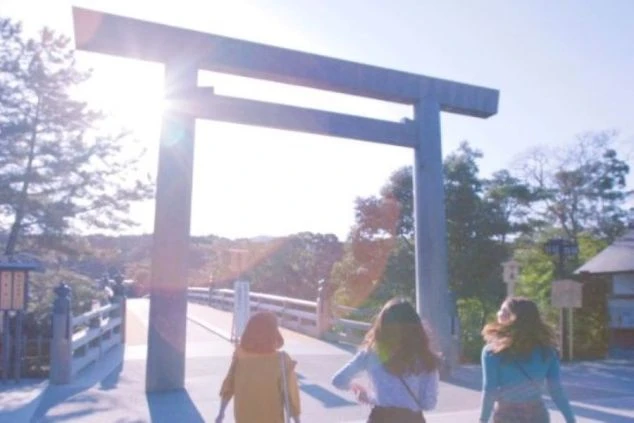 夕暮れ時、木製の鳥居をくぐり橋を渡る3人の女性の後ろ姿。逆光で光が差し込み、厳かで神聖な場所の雰囲気。リゾートバイトの休日に観光を楽しむイメージ。