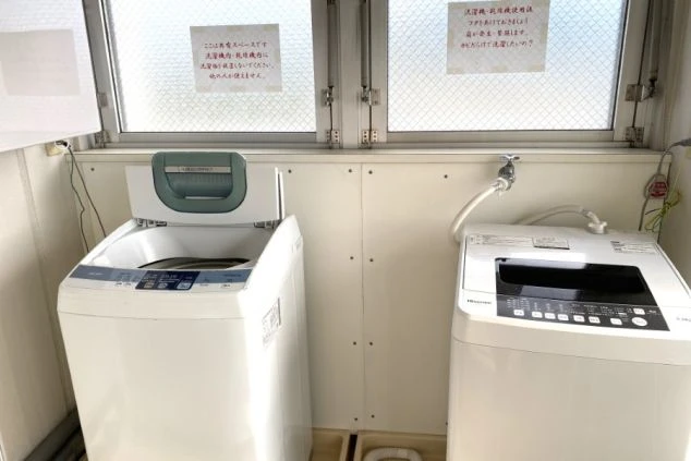 2台の洗濯機が並べられたランドリースペースの画像。窓から光が差し込み、壁には洗濯に関する注意書きが貼られている。リゾートバイトの寮の設備イメージ。