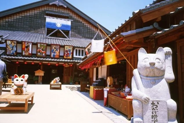 伊勢神宮「おかげ横丁」の入り口付近の画像。大きな招き猫の石像があり、和風の建物には暖簾や提灯が飾られている。活気ある観光地のイメージ。