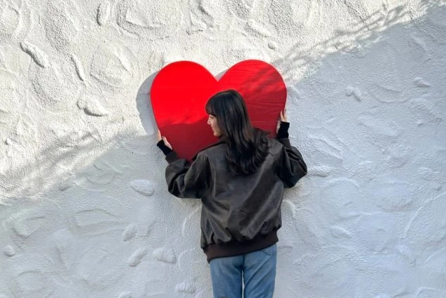 白い漆喰壁に設置された大きな赤いハートのオブジェを、両手で抱えるようにして立つ女性の背中。