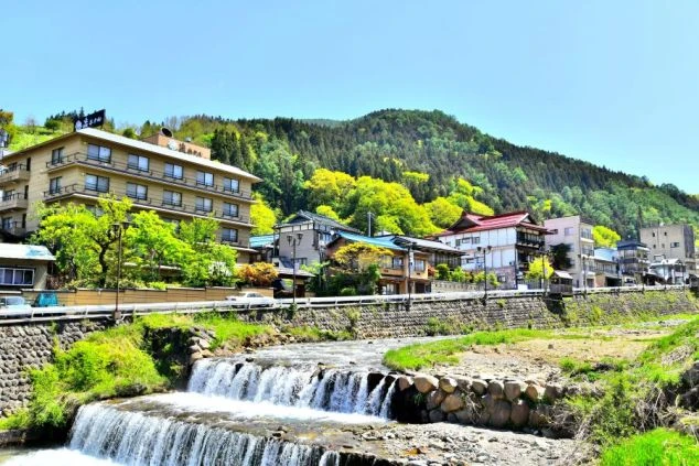緑豊かな山を背景に、川沿いに旅館や建物が立ち並ぶ温泉街の全景画像。日本の情緒あふれる温泉地でのゆったりとした滞在を想像させる。