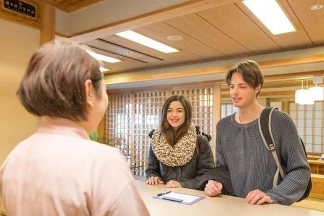 旅館のフロントで、笑顔の女性スタッフと外国人観光客の男女がチェックインをしている画像。日本の温泉旅館で旅の始まりを感じる。