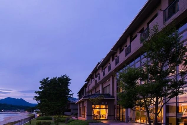 夕暮れのホテル外観。ビーチ沿いに建ち、室内から光が漏れる職場のイメージ画像。