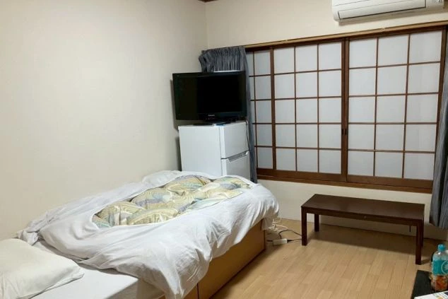 寮の個室。ベッド、冷蔵庫、テレビ、エアコン、障子付きの部屋が映る。