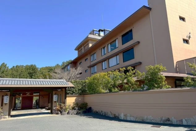 緑に囲まれた和風の建物。茶色い壁が特徴的で、手前には駐車場と入口が見えます。