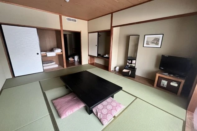 リゾートバイトの客室寮の和室の部屋の画像。黒い座卓とピンク色の座布団が置かれ、テレビとドレッサーが備え付けられている。収納スペースもあり、広々とした部屋の画像。