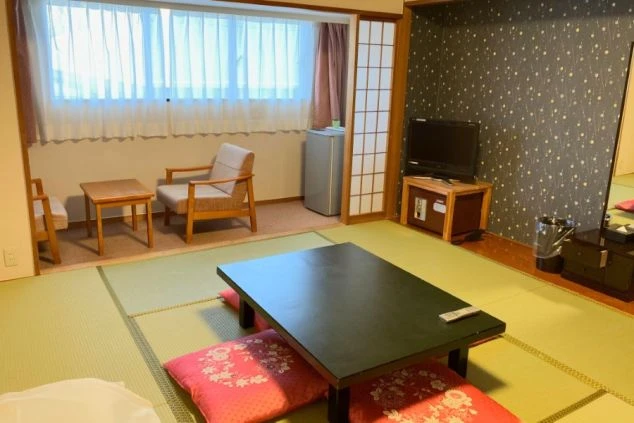 リゾートバイトの客室寮の和室の部屋の画像。黒い座卓とピンク色の座布団が置かれ、テレビとドレッサーが備え付けられている。収納スペースもあり、広々とした部屋の画像。