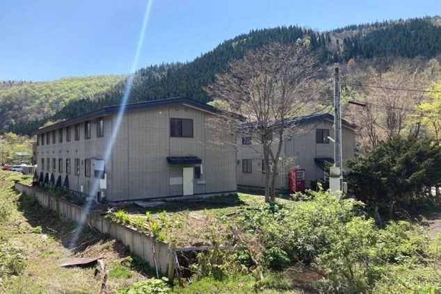 寮の外観。緑の山を背景に、2棟の建物と自動販売機が映る。