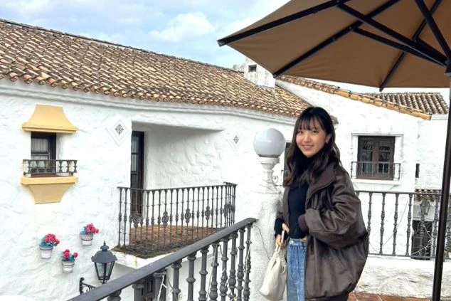 白い壁と瓦屋根の異国情緒あふれる建物を背景に、笑顔で立つ女性。リゾート地での楽しそうな様子が伝わる。