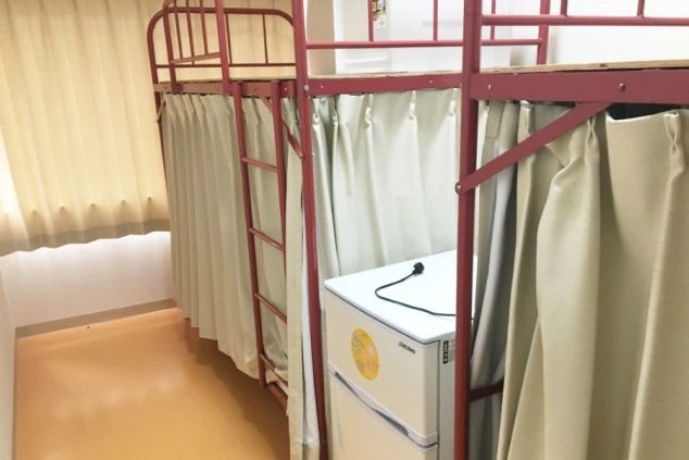 赤色の二段ベッドが並んだ寮の画像です。ベッドの間に冷蔵庫が設置されており、各個人のスペースに家電が備え付けられているリゾートバイト寮のイメージです。