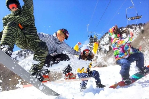 夕暮れの雪山ゲレンデで、スノーボードウェアを着た人が両手を上げて雪を撒き散らしている画像です。地平線に沈む太陽が輝き、リゾートバイトの休日に夕焼けの雪山を楽しむ様子が伝わります。