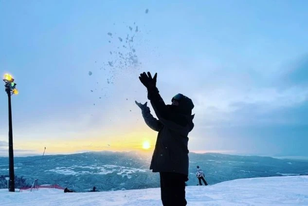 夕暮れの雪山ゲレンデで、スノーボードウェアを着た人が両手を上げて雪を撒き散らしている画像。地平線に沈む太陽が輝き、仕事の合間に夕焼けの雪山を楽しむ様子が伝わります。