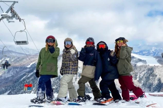 雪山ゲレンデに立つ5人の女性が、スノーボードウェアを着て笑顔でポーズを取っている画像です。遠くにはリフトも見え、リゾートバイトの休日に仲間とスノボを楽しむ様子が伝わります。
