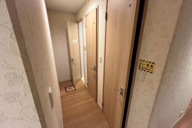 ドアがいくつかある廊下。片方のドアは開いており、小さな部屋が見えている。壁には花柄の壁紙が貼られている。