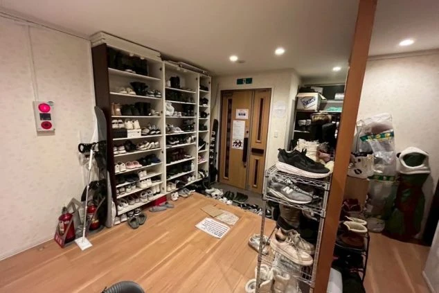 多数の靴が棚にきれいに並べられた部屋の入り口。床にはいくつかの靴とスノーボード、消火器、そして整理されていない用具の山がある。