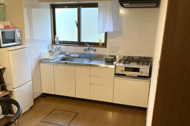 リゾートバイトの寮のキッチンの画像。冷蔵庫、電子レンジ、シンク、ガスコンロが備わったシステムキッチンで、窓からは光が差し込む。自炊に便利な設備が整っている。