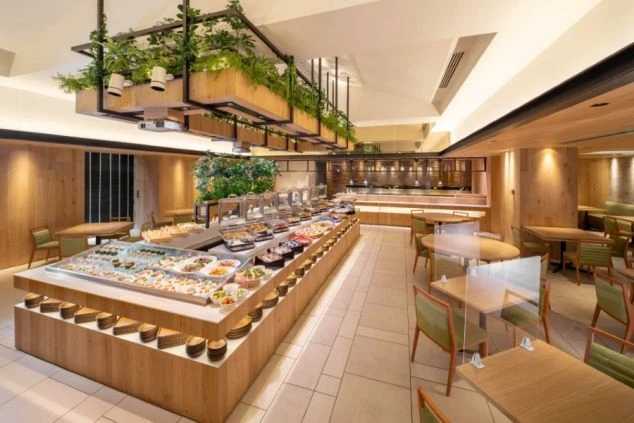 ホテルレストランの豪華なビュッフェ台。彩り豊かな料理が並び、客席と調理場が映る職場のイメージ画像。