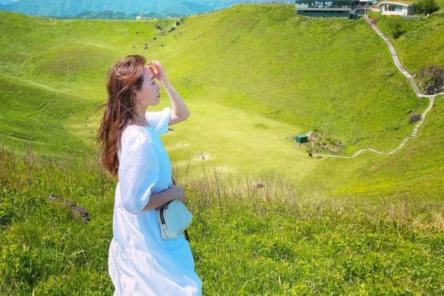 緑の高原に立つ女性の画像。白いワンピースを着用し、手で日差しを遮りながら遠くを眺めている。リゾートバイトの休日に楽しめる、開放的な自然風景。