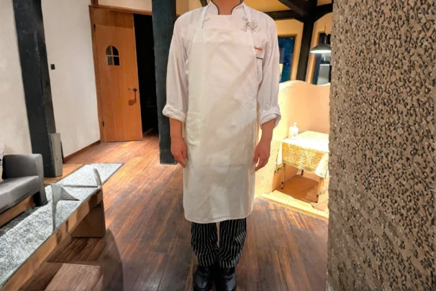 白いコックコートとエプロン、ストライプのパンツを着用し、調理場またはレストランのような場所で直立している人物。