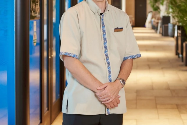 男性スタッフの制服。ベージュの開襟シャツの襟元と袖口に、沖縄の伝統的な柄のような青い模様のアクセントが入っている。