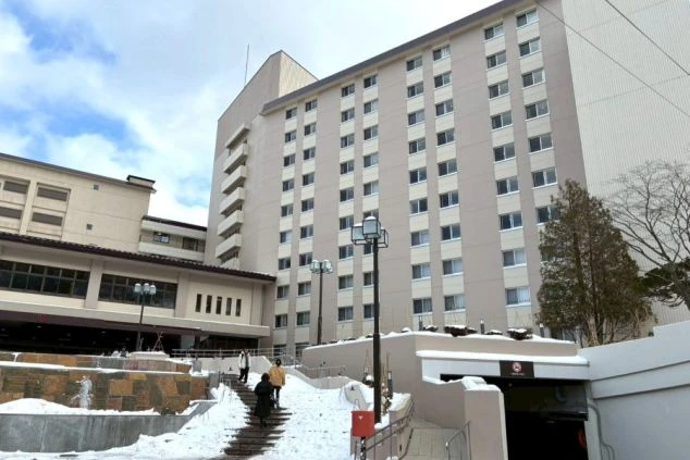 登別地獄谷の看板の前で笑顔で立つ2人の女性。背景には荒々しい火山地形と緑の山々が広がる。リゾートバイトの休日の過ごし方のイメージ。