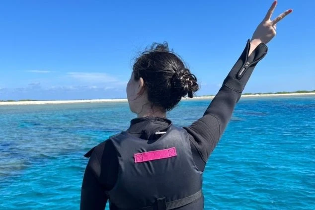 沖縄の海で、青い空青い海を背景に20代女性リゾートバイトスタッフが背中を向けピースする様子のワクトリオリジナル画像。