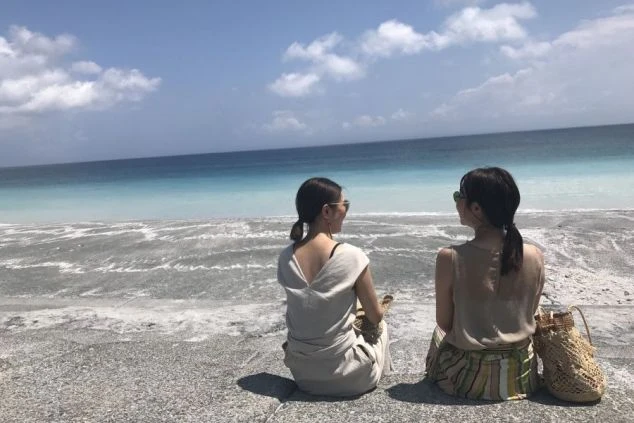 海辺に座って話す2人の女性の後ろ姿の画像。青いグラデーションの海が広がり、リゾートバイトの休日に友人との時間を楽しんでいる様子。