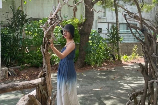 青と白のグラデーションワンピースを着た女性が、木のオブジェに手を添えて自然の中で佇む様子。リゾートバイトの休日を満喫し、自然に囲まれた場所でリラックスしたい方に魅力が伝わる画像。