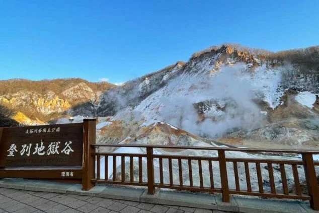 雪景色と噴煙が上がる登別地獄谷の絶景。手前には「登別地獄谷」と書かれた看板と柵が見える。