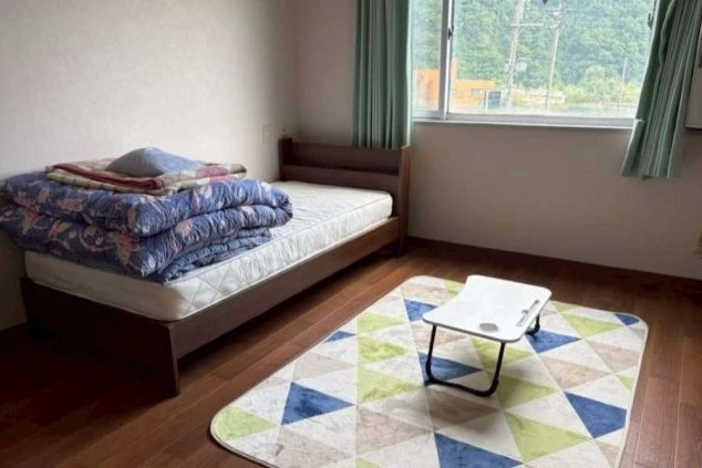 リゾートバイトの個室寮の室内。ベッド、テーブル、ラグが敷かれており、窓からは豊かな自然が見える。プライベートな空間が確保され、快適な生活を送れることをアピール。