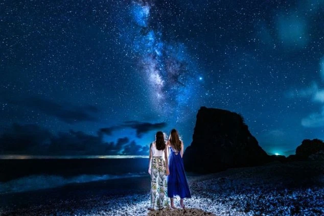 満天の星空と天の川が輝く夜のビーチに立つ2人の女性。ロマンチックなリゾートの夜景。