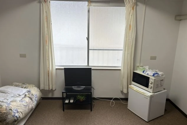 シンプルな寮の個室。シングルベッドの横に、テレビ、小型冷蔵庫、その上に電子レンジがコンパクトに配置されている。窓には白いカーテンがかかり、落ち着いた雰囲気。