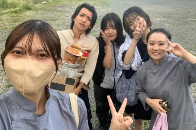 屋外で若い男女5人が笑顔で写っている自撮り画像です。手前の女性はマスクをしており、その横の男性はカップ麺を抱えています。リゾートバイトの仲間との集合写真で、楽しい雰囲気が伝わります。