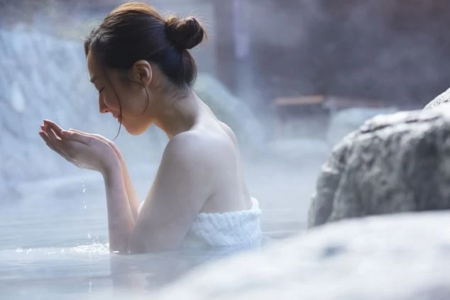 湯気が立ち込める露天風呂で、湯をすくって顔に当てる女性。リゾートバイトの疲れを癒す温泉のイメージ。