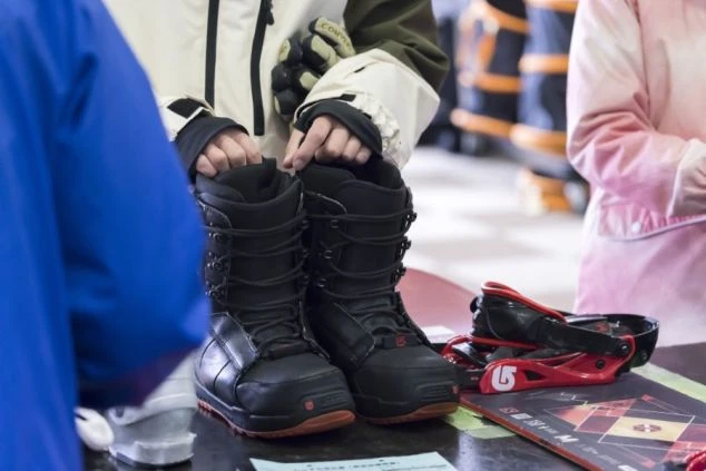 スノーボードブーツを持つ手元と、スノーボード用品が並べられた様子。スキー場のレンタルショップの接客イメージ。