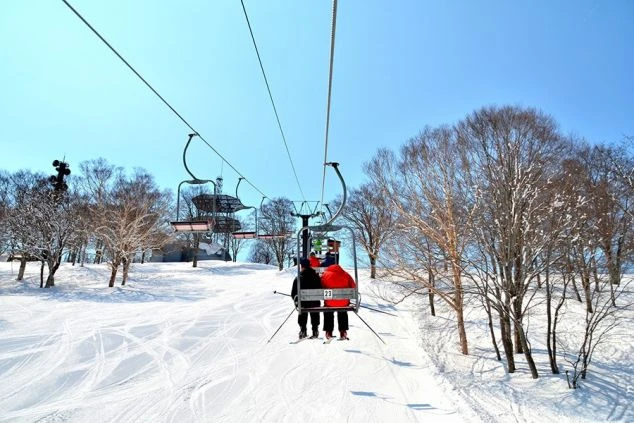 快晴のスキー場で、リフトに乗ってゲレンデを上るスキーヤーたち。雪景色と木々が広がる。