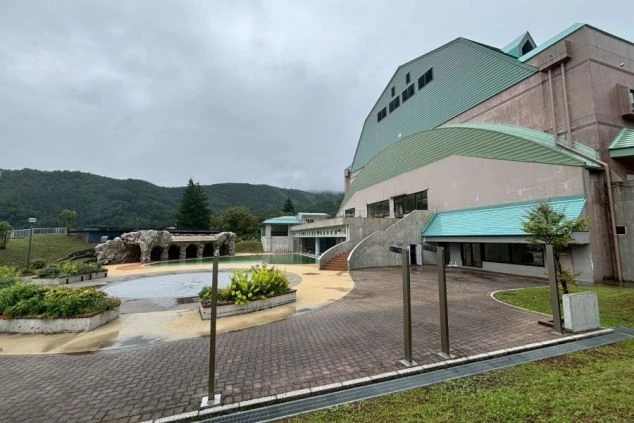 野沢温泉スパリーナの、プールや広場が併設された温泉施設の外観。曇り空の下で、建物の緑色の屋根が目立つ。