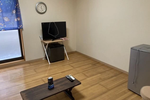 寮の個室。フローリングの床に、テレビ台、小型冷蔵庫、壁掛け時計、エアコンが設置されたシンプルな和モダンの空間です。