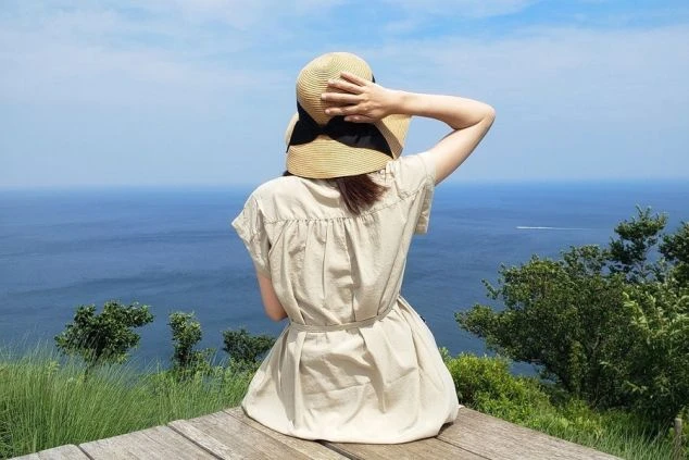 海を見下ろす高台で、麦わら帽子を被って座る女性の後ろ姿。広がる青い海と空が開放的なリゾートの景色。