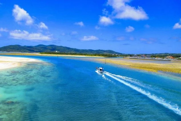 ターコイズブルーの美しい海を船が航行する、沖縄県久米島の風景。手前には白い砂浜と、奥には緑豊かな島々が広がり、マリンアクティビティやビーチでのリゾートバイトに興味がある方に魅力的な画像。