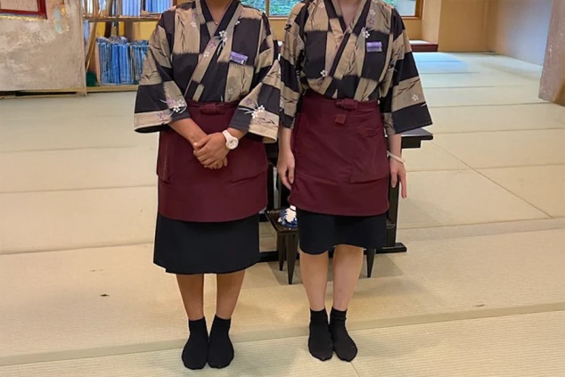 ベージュと黒の和風柄の作務衣に、エンジ色のエプロンを着用。和の雰囲気を伝える旅館接客の制服。
