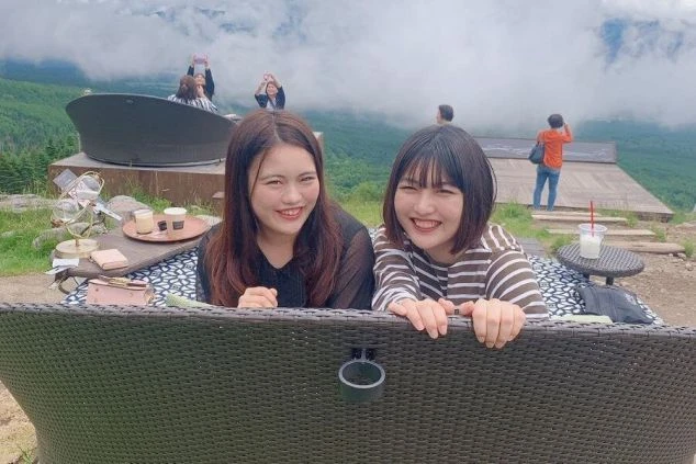 リゾート地の展望テラスで、笑顔でポーズをとる2人の女性。奥には景色を楽しむ人々が見える。リゾートバイトの休日に派遣仲間と過ごすイメージ。