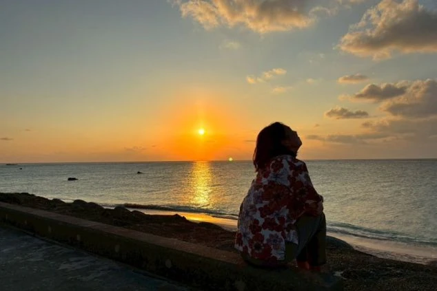 沖縄県宮古島の海岸沿いで、水平線に沈む黄金色の夕日を静かに見つめる女性の後ろ姿の画像。空と海がオレンジ色に溶け合う絶景の中、心地よい海風を感じながらリラックスする休日のひととき。