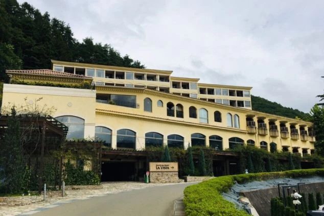 南欧風の明るい黄色の外壁と、アーチ状の窓が特徴的なリゾートホテル。手前に車寄せへ続く坂道と生垣が見える。