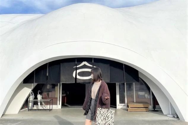 曲線が特徴的な白い建物の前で、ゼブラ柄のバッグを持ちポーズをとる女性。和風の暖簾がかかっている。