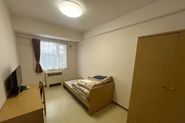 寮の個室。ベッド、机、テレビ、冷蔵庫、クローゼット、暖房器具が映る。